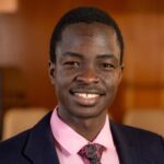 Photo of man wearing black blazer, tie, and pink collared shirt. Man is smiling.